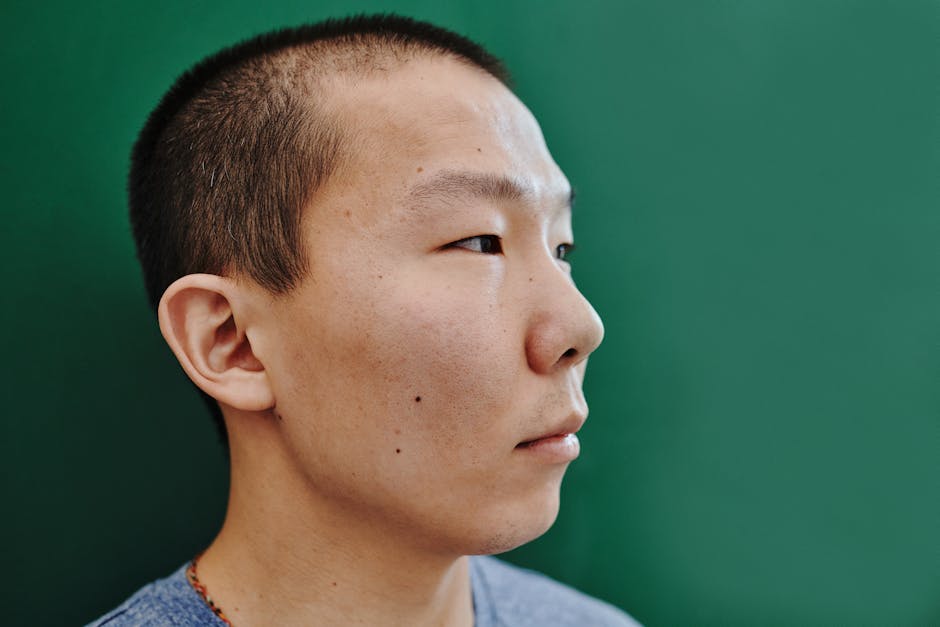 Close-up profile of an East Asian man against a green background, showcasing short hair.