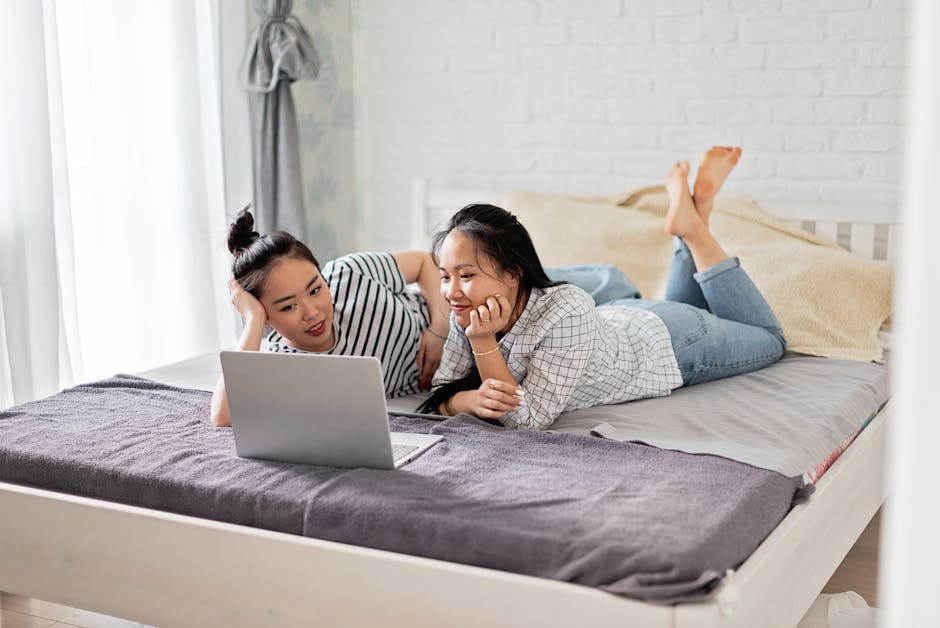 Two women lying on a bed, enjoying a laptop in a cozy bedroom setting. Ideal for lifestyle and technology themes.