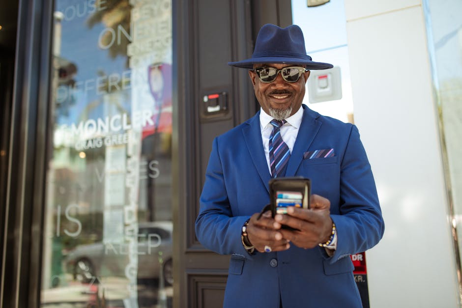 A fashionable man in a blue suit and hat smiles while checking his smartphone outside a store.