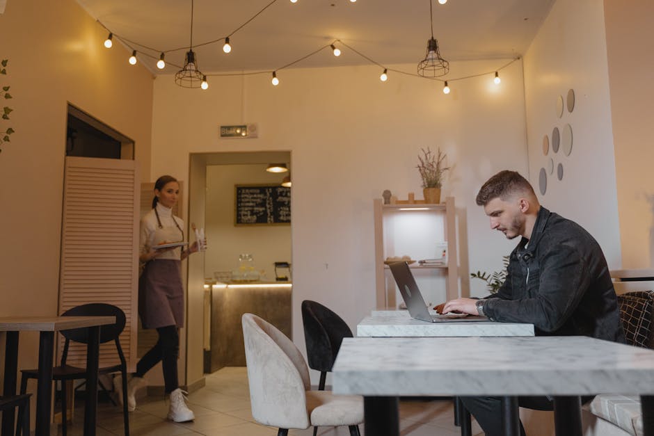 Man working on laptop in a stylish café while a waitress serves customers.