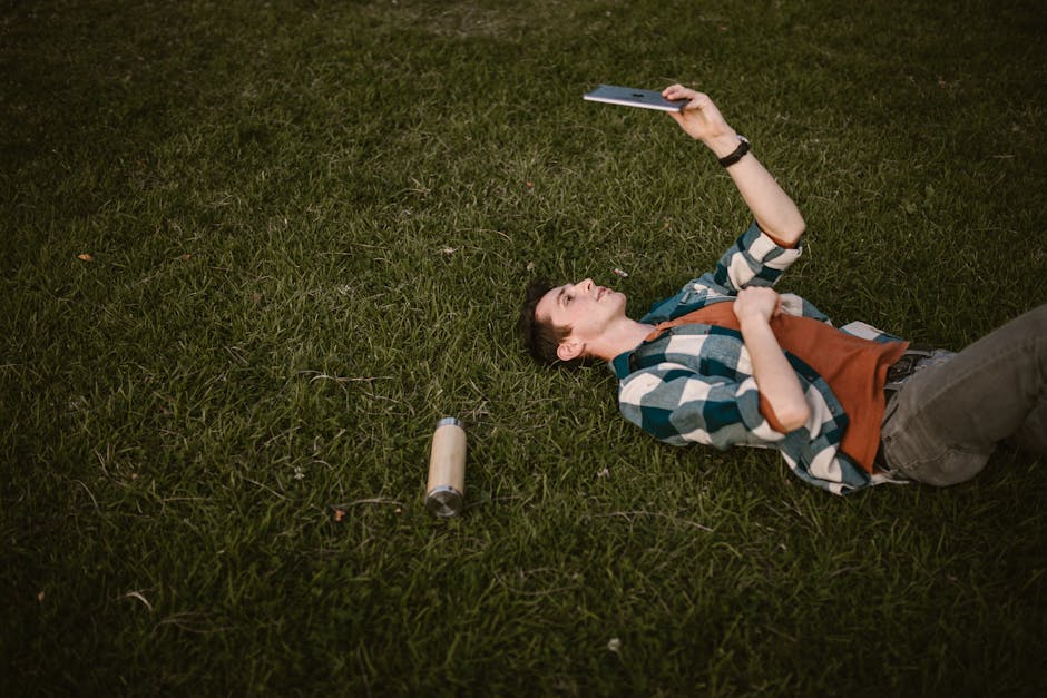 Male lying in park using tablet, green grass background, casual attire.