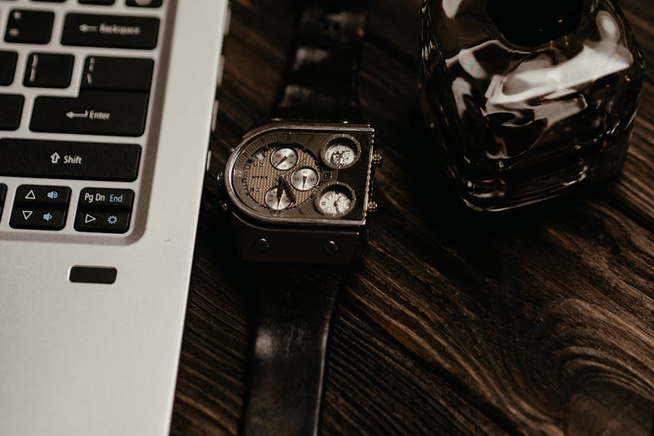Wooden table featuring a laptop, wristwatch, and perfume bottle in a moody setting.