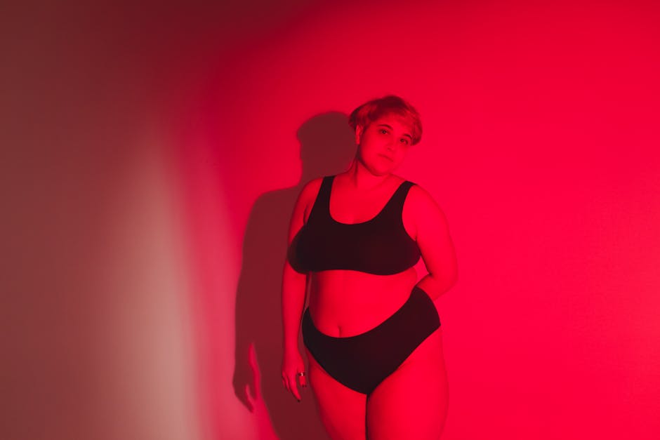 A confident woman poses in red lighting, celebrating body positivity in a studio setting.