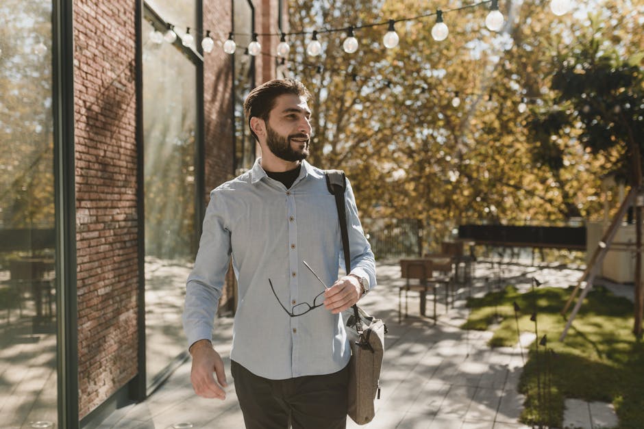 A smiling man in blue long sleeves walking outdoors in a casual setting, holding eyeglasses.