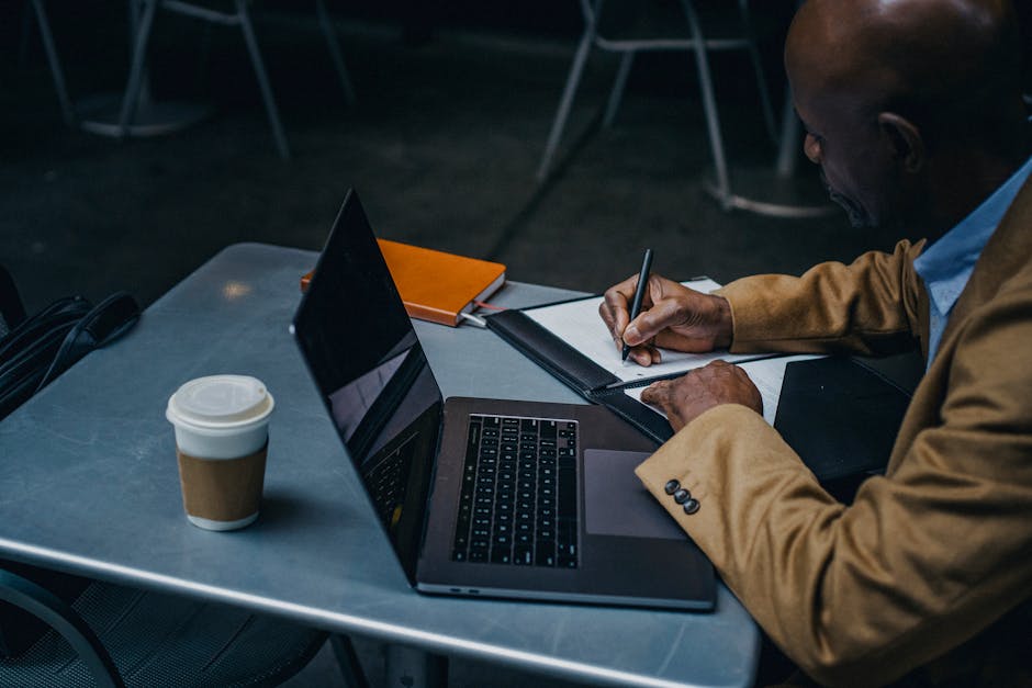 From above of focused black freelancer sitting at table with laptop and writing notes on paper