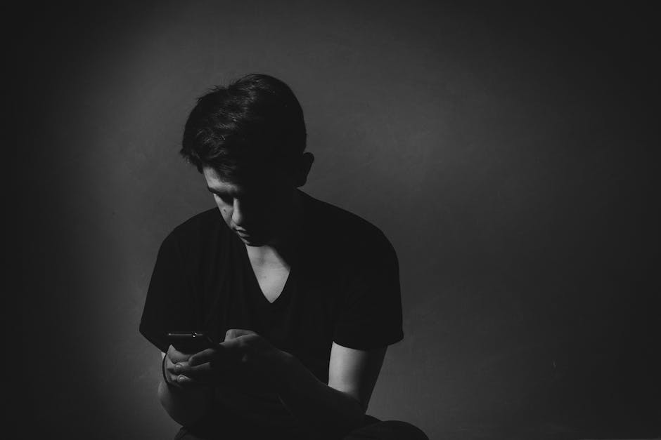 A young man in a dark room, focused on his smartphone. Black and white portrait captures a thoughtful mood.